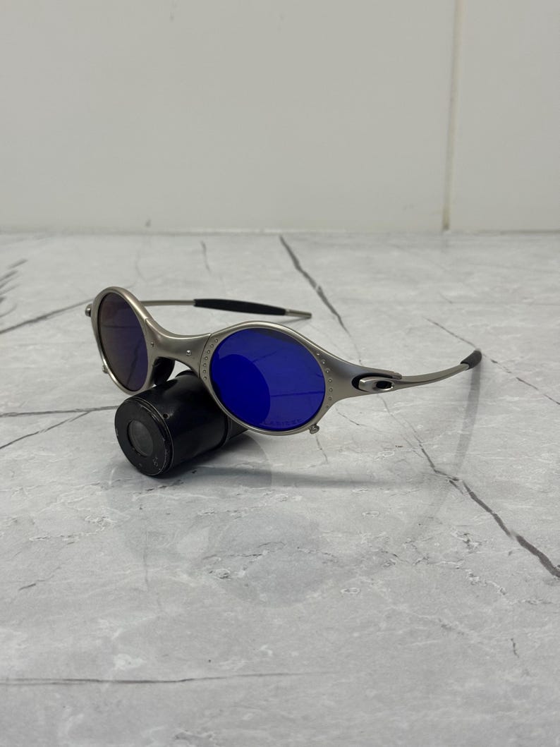 May include: Silver-framed sunglasses with blue lenses rest on a black cylindrical case. The sunglasses have a sleek, modern design with a curved frame and dark temples. The lenses are a vibrant blue color.