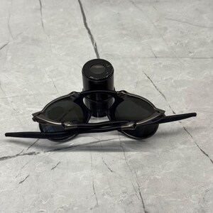 以下が含まれることがあります： 黒いサングラス。濃い色のレンズと洗練されたデザイン。サングラスは黒いフレームとテンプルがあり、黒い円筒形の物体の上に置かれています。レンズは着色されているように見え、太陽からの保護を提供します。