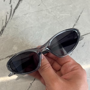 May include: Gray sunglasses with dark lenses. The sunglasses have a sleek, oval shape and a transparent frame. The arms are thin and extend to the temples. The sunglasses are held by a hand.