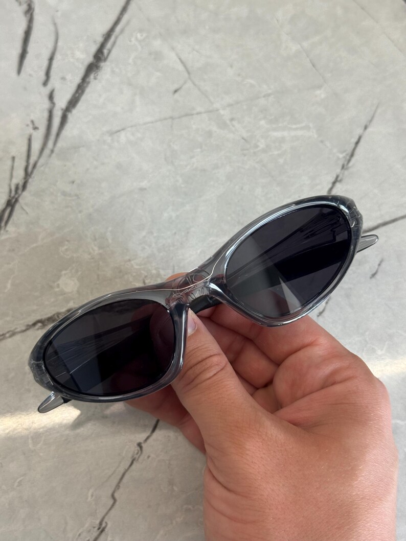 May include: Gray sunglasses with dark lenses. The sunglasses have a sleek, oval shape and a transparent frame. The arms are thin and extend to the temples. The sunglasses are held by a hand.