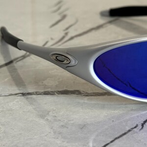 May include: Close-up of silver sunglasses with blue lenses. The sunglasses have a sleek, modern design with a curved frame and dark grey ear stems. The lenses are a vibrant shade of blue.
