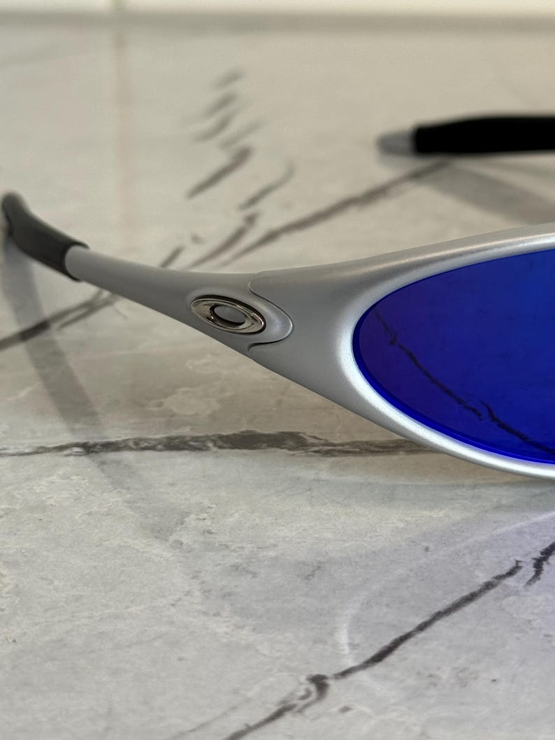 May include: Close-up of silver sunglasses with blue lenses. The sunglasses have a sleek, modern design with a curved frame and dark grey ear stems. The lenses are a vibrant shade of blue.