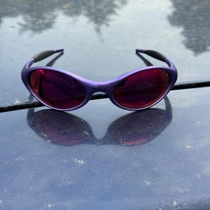May include: Purple sunglasses with dark red lenses. The sunglasses have a rounded frame and black accents on the temples. The lenses reflect the surrounding environment. The sunglasses are resting on a dark surface.