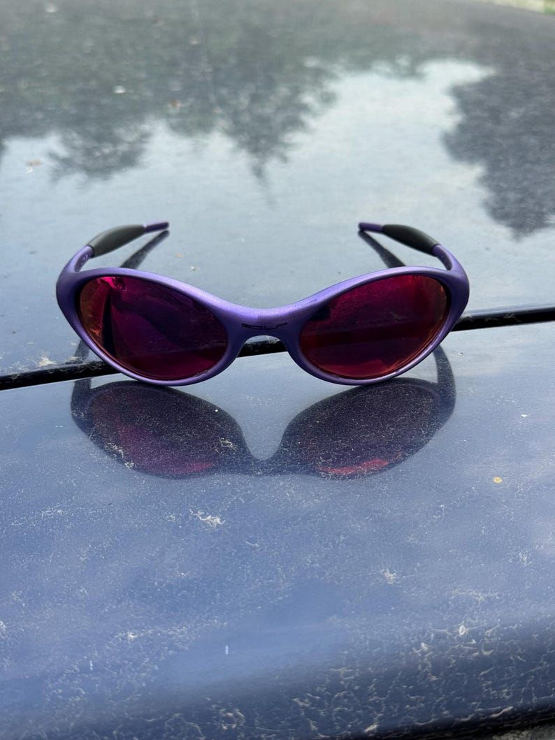 May include: Purple sunglasses with dark red lenses. The sunglasses have a rounded frame and black accents on the temples. The lenses reflect the surrounding environment. The sunglasses are resting on a dark surface.