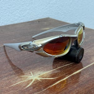 Puede incluir: Gafas de sol plateadas y doradas con lentes naranjas y amarillas. Las gafas de sol tienen un dise&ntilde;o elegante y moderno con una montura curvada y un peque&ntilde;o objeto cil&iacute;ndrico negro adjunto en la parte inferior.