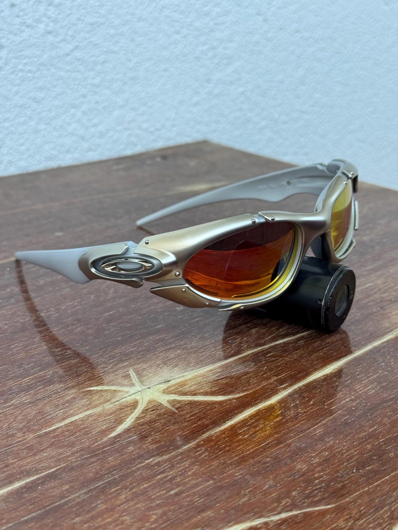 Puede incluir: Gafas de sol plateadas y doradas con lentes naranjas y amarillas. Las gafas de sol tienen un dise&ntilde;o elegante y moderno con una montura curvada y un peque&ntilde;o objeto cil&iacute;ndrico negro adjunto en la parte inferior.