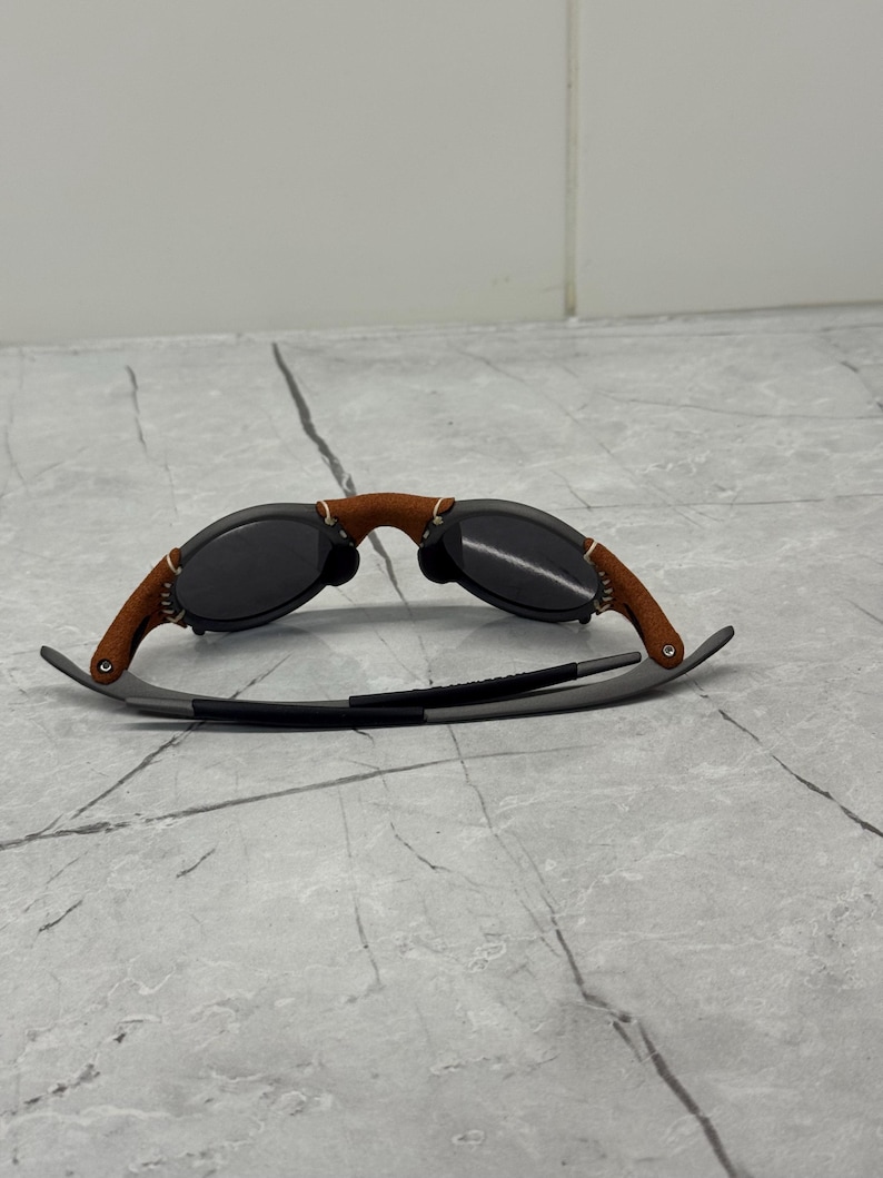 May include: Sunglasses with a brown leather bridge and temples, and dark lenses. The frames are a combination of gray metal and brown leather. The sunglasses are designed for eye protection and fashion.