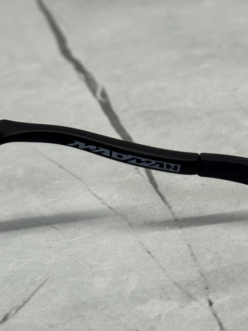 May include: Black eyeglasses frame with the word "MADMAN" printed in white on the temple. The frame is set against a light grey and white background.