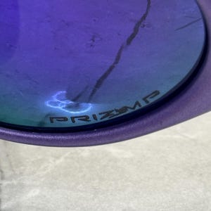 May include: Close-up of a purple-framed sunglass lens with a blue-tinted surface. The word "PRIZM" is printed on the lens. A crack is visible on the lens.