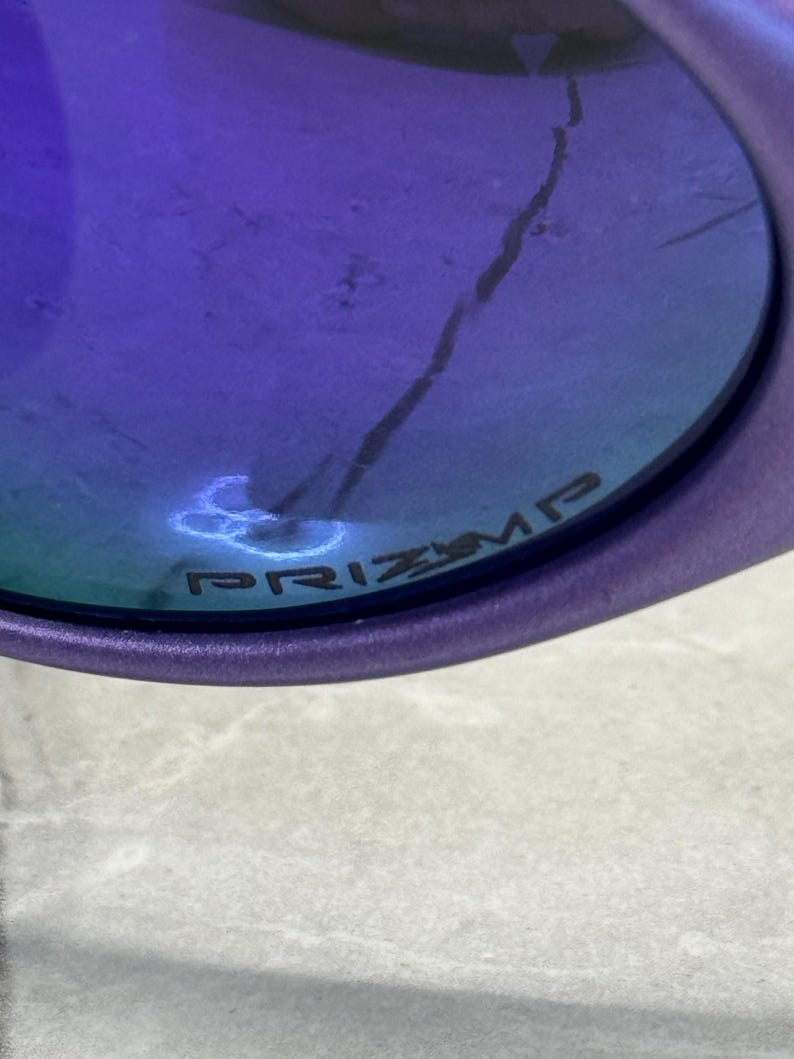 May include: Close-up of a purple-framed sunglass lens with a blue-tinted surface. The word "PRIZM" is printed on the lens. A crack is visible on the lens.