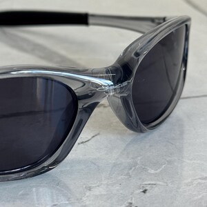 May include: Close-up of gray sunglasses with dark lenses. The sunglasses have a sleek, modern design with a curved frame and a black strap. The lenses are dark, providing shade and protection from the sun.