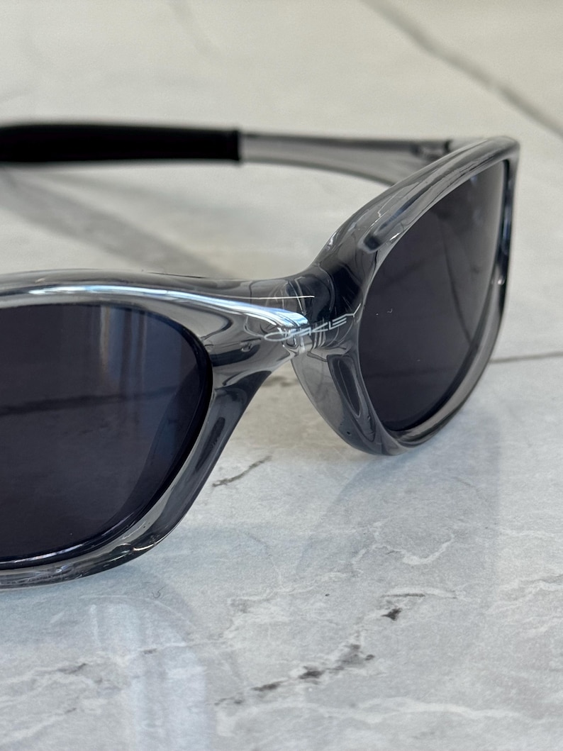May include: Close-up of gray sunglasses with dark lenses. The sunglasses have a sleek, modern design with a curved frame and a black strap. The lenses are dark, providing shade and protection from the sun.