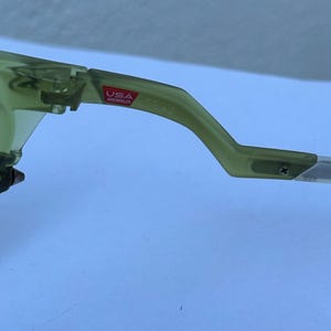 May include: Close-up of a pair of olive green sunglasses. The frame is a translucent green with a red "USA Assembled" sticker on the arm. The arm is curved and attached to the lens.
