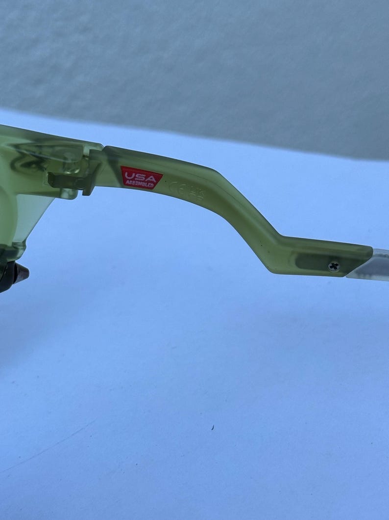 May include: Close-up of a pair of olive green sunglasses. The frame is a translucent green with a red "USA Assembled" sticker on the arm. The arm is curved and attached to the lens.