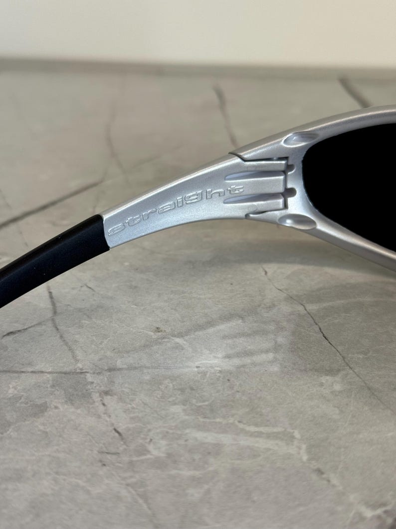 May include: Close-up of silver sunglasses with black lenses. The word "straight" is printed on the silver frame. The sunglasses have a sleek, modern design.