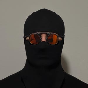 Pu&ograve; includere: Passamontagna nero con occhiali da sole con lenti arancioni. Gli occhiali da sole hanno una montatura scura e lenti rotonde. Il passamontagna copre la testa e il collo, con aperture per gli occhi.