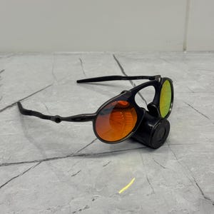 Pu&ograve; includere: Occhiali da sole neri con lenti arancioni e verdi. Gli occhiali hanno un design unico con lenti rotonde e una montatura nera. Le lenti presentano un effetto sfumato, che passa dall'arancione al verde. Progettati per l'uso all'aperto.
