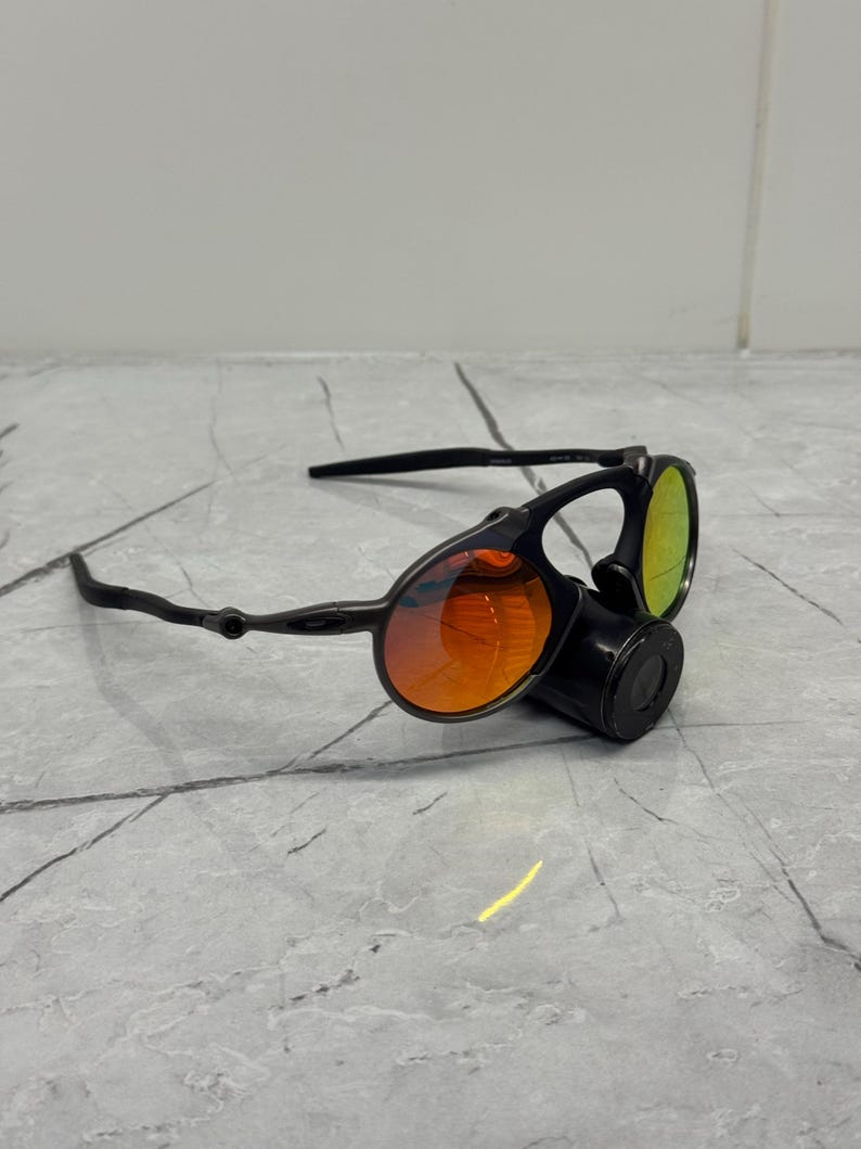 Pu&ograve; includere: Occhiali da sole neri con lenti arancioni e verdi. Gli occhiali hanno un design unico con lenti rotonde e una montatura nera. Le lenti presentano un effetto sfumato, che passa dall'arancione al verde. Progettati per l'uso all'aperto.