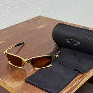 May include: Gold-framed sunglasses with brown lenses, a black case, and a black cleaning cloth. The sunglasses have a rectangular shape and a sleek, modern design. The case and cloth feature the Oakley logo.