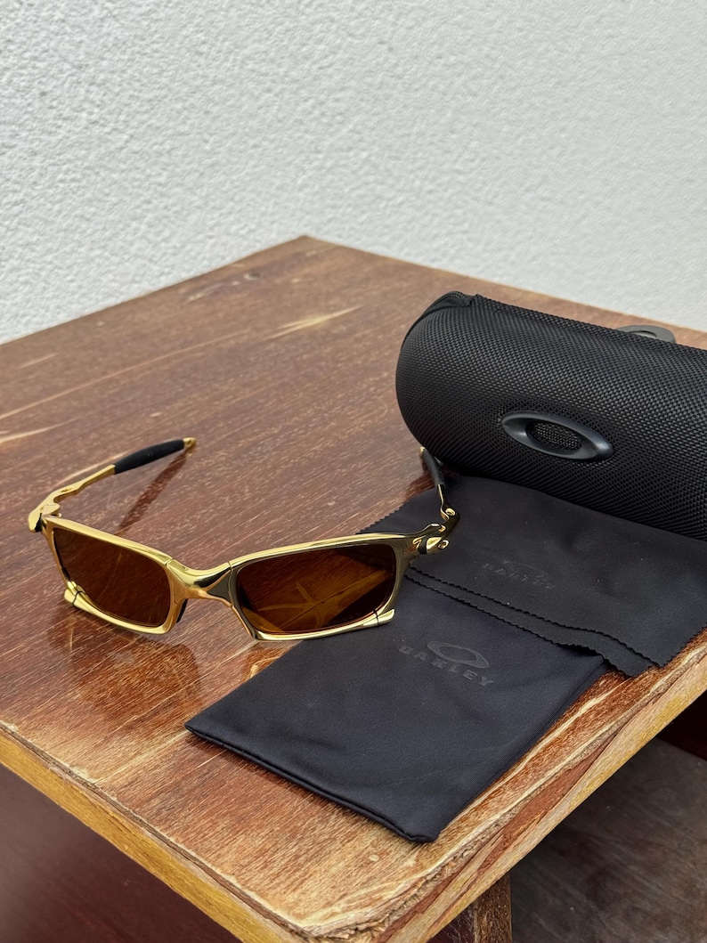 May include: Gold-framed sunglasses with brown lenses, a black case, and a black cleaning cloth. The sunglasses have a rectangular shape and a sleek, modern design. The case and cloth feature the Oakley logo.