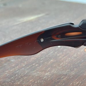 Puede incluir: Primer plano de una patilla de gafas de sol con estampado marr&oacute;n y negro. La patilla tiene el texto "J-A" y un peque&ntilde;o detalle plateado. Las gafas de sol est&aacute;n sobre una superficie de madera.