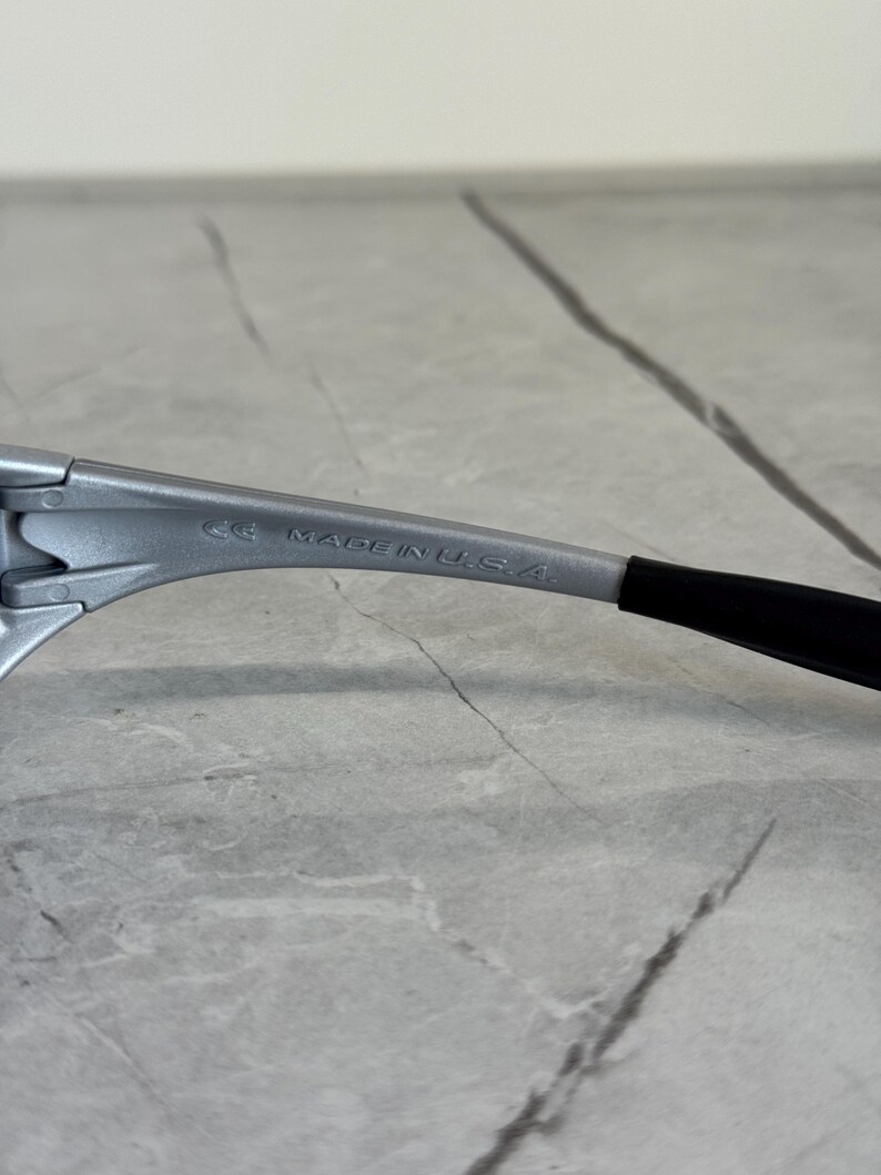 Puede incluir: Primer plano de una montura de gafas plateada con una patilla negra. El texto "MADE IN U.S.A." est&aacute; impreso en la montura.