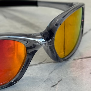 K&ouml;nnte beinhalten: Nahaufnahme einer Sonnenbrille mit einem durchscheinenden grauen Rahmen und verspiegelten Gl&auml;sern, die von Rot nach Gelb &uuml;bergehen. Die Sonnenbrille hat ein schlankes, modernes Design, wobei der Markenname auf dem Rahmen sichtbar ist.