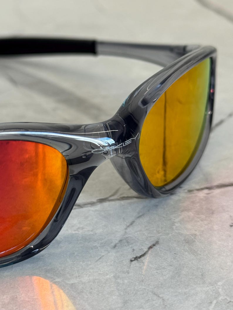 K&ouml;nnte beinhalten: Nahaufnahme einer Sonnenbrille mit einem durchscheinenden grauen Rahmen und verspiegelten Gl&auml;sern, die von Rot nach Gelb &uuml;bergehen. Die Sonnenbrille hat ein schlankes, modernes Design, wobei der Markenname auf dem Rahmen sichtbar ist.