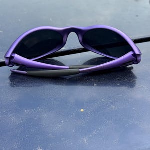 May include: Purple sunglasses with black lenses. The sunglasses have a sleek, modern design with a matte finish. The frame is a vibrant purple color, and the lenses are dark, providing shade and protection from the sun.