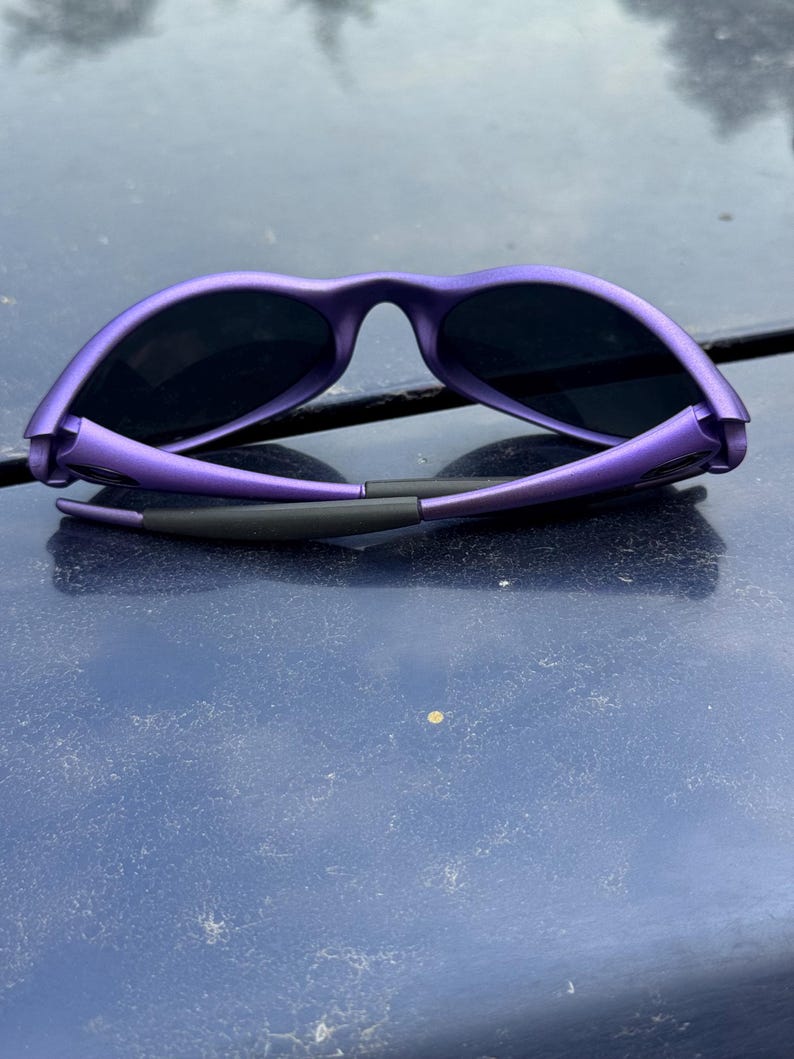 May include: Purple sunglasses with black lenses. The sunglasses have a sleek, modern design with a matte finish. The frame is a vibrant purple color, and the lenses are dark, providing shade and protection from the sun.
