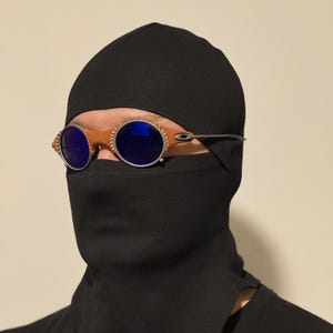 May include: Round, brown-rimmed sunglasses with blue lenses. The sunglasses are worn with a black head covering and face mask. The frame has a silver metal arm.