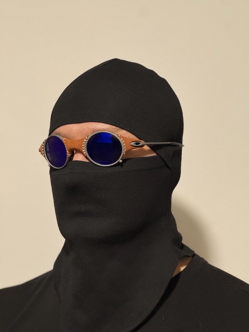 May include: Round, brown-rimmed sunglasses with blue lenses. The sunglasses are worn with a black head covering and face mask. The frame has a silver metal arm.