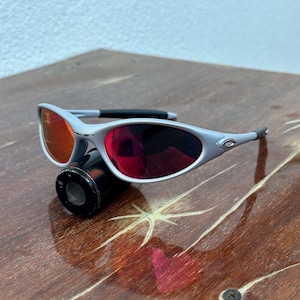 May include: Silver-framed sunglasses with red-tinted lenses. The sunglasses have a sleek, modern design with a black nose bridge and temples. The lenses are a vibrant red color, and the frame is a light silver color.
