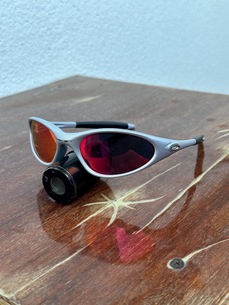 May include: Silver-framed sunglasses with red-tinted lenses. The sunglasses have a sleek, modern design with a black nose bridge and temples. The lenses are a vibrant red color, and the frame is a light silver color.