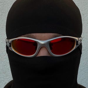May include: Silver-framed sunglasses with red lenses are worn over a black balaclava. The sunglasses have a curved design and are set against a neutral background. The overall aesthetic is modern and edgy.