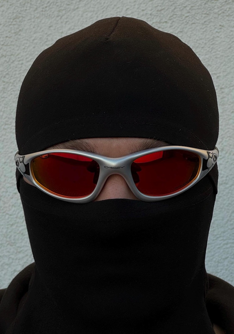 May include: Silver-framed sunglasses with red lenses are worn over a black balaclava. The sunglasses have a curved design and are set against a neutral background. The overall aesthetic is modern and edgy.