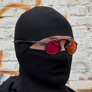 May include: Black balaclava and sunglasses with red lenses. The sunglasses have a black frame and are worn over the balaclava. The background is a weathered brick wall.