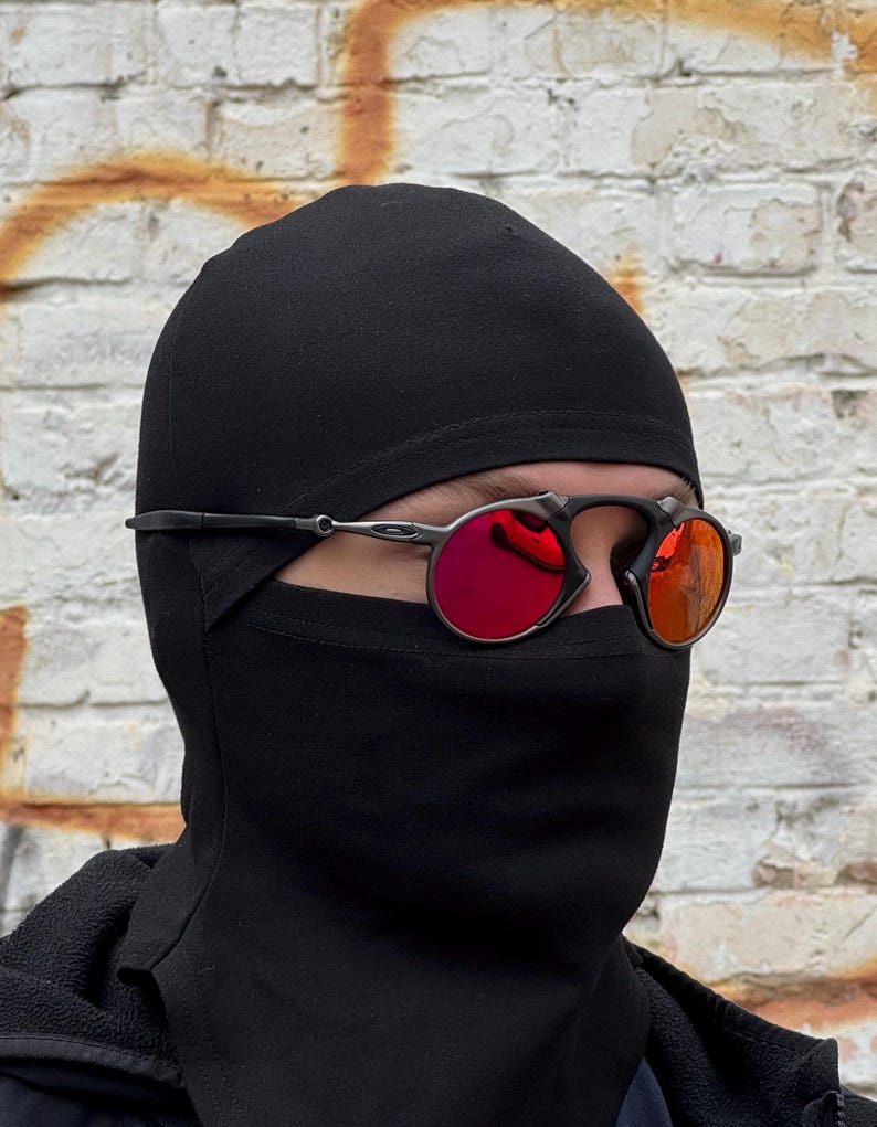 May include: Black balaclava and sunglasses with red lenses. The sunglasses have a black frame and are worn over the balaclava. The background is a weathered brick wall.