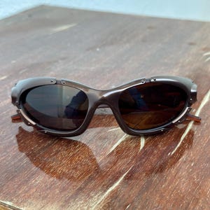 Puede incluir: Gafas de sol marrones con un dise&ntilde;o de montura curvada &uacute;nico. Las lentes son oscuras y la montura tiene peque&ntilde;os detalles met&aacute;licos. Las patillas de las gafas son de color marr&oacute;n claro. Est&aacute;n sobre una superficie de madera.