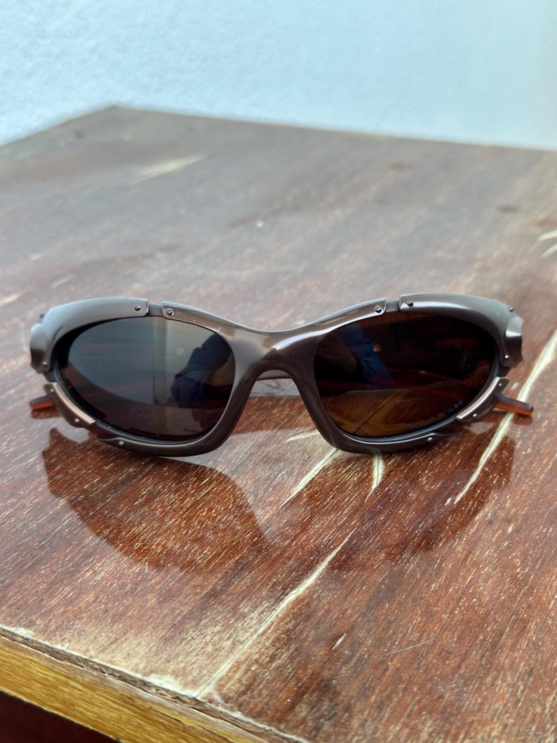 Puede incluir: Gafas de sol marrones con un dise&ntilde;o de montura curvada &uacute;nico. Las lentes son oscuras y la montura tiene peque&ntilde;os detalles met&aacute;licos. Las patillas de las gafas son de color marr&oacute;n claro. Est&aacute;n sobre una superficie de madera.