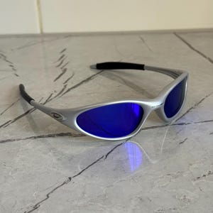 May include: Silver sunglasses with blue lenses. The frame has a curved design with black temple tips. These sunglasses offer a modern look. Suitable for outdoor activities, they provide both style and eye protection.