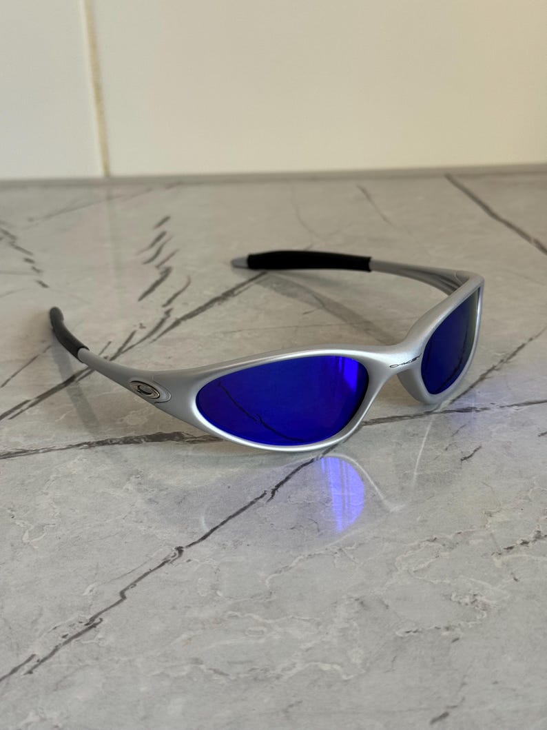 May include: Silver sunglasses with blue lenses. The frame has a curved design with black temple tips. These sunglasses offer a modern look. Suitable for outdoor activities, they provide both style and eye protection.
