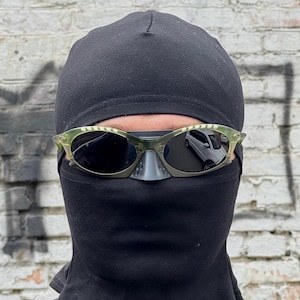 May include: A person wearing a black balaclava and olive green sunglasses. The sunglasses have dark lenses and a unique frame design. The background is a weathered brick wall.