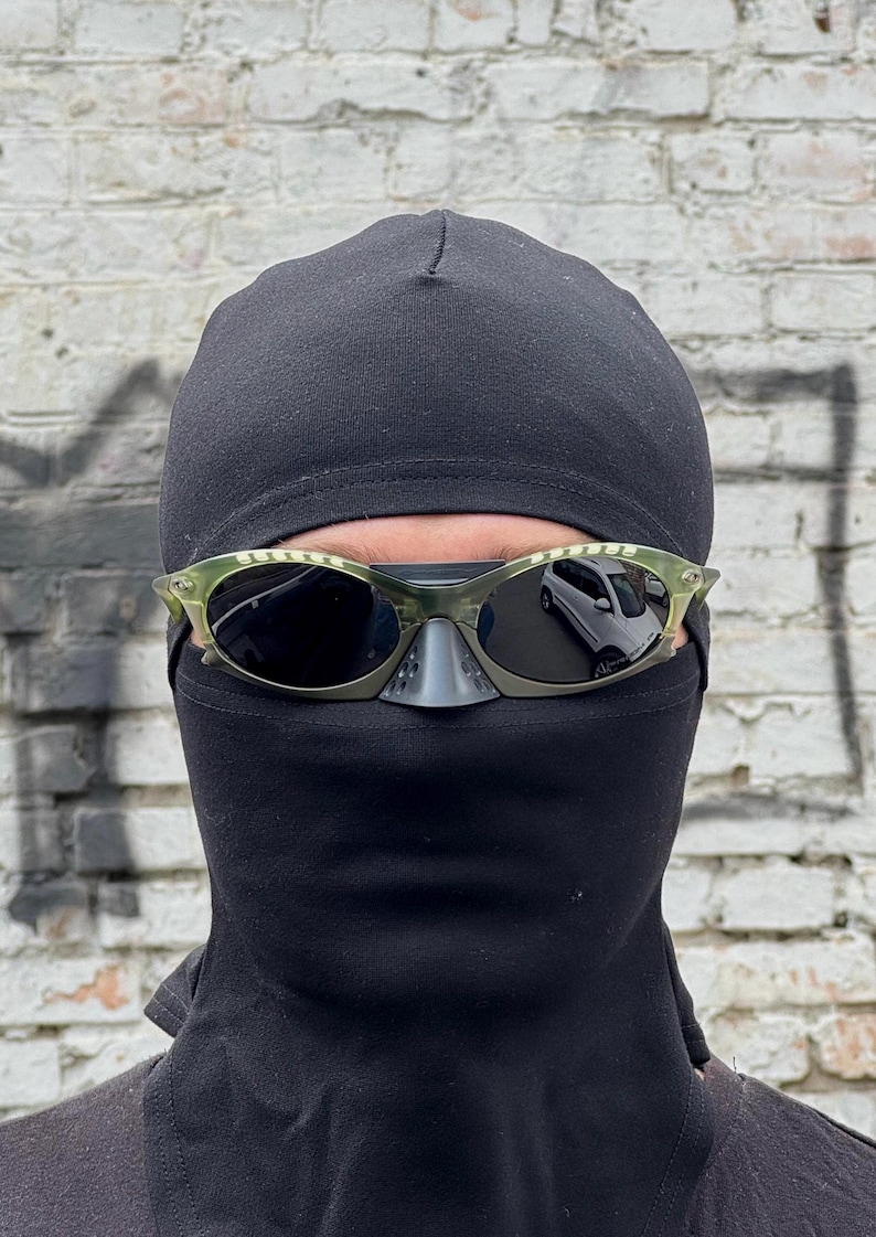 May include: A person wearing a black balaclava and olive green sunglasses. The sunglasses have dark lenses and a unique frame design. The background is a weathered brick wall.