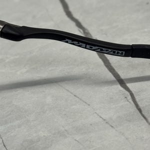 Pu&ograve; includere: Primo piano di un'asta di occhiali nera con la scritta "MADMAN" stampata in argento. L'asta &egrave; curva e ha una finitura opaca. Lo sfondo &egrave; una superficie grigio chiaro con linee grigio scuro.
