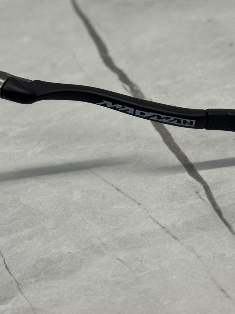 Pu&ograve; includere: Primo piano di un'asta di occhiali nera con la scritta "MADMAN" stampata in argento. L'asta &egrave; curva e ha una finitura opaca. Lo sfondo &egrave; una superficie grigio chiaro con linee grigio scuro.