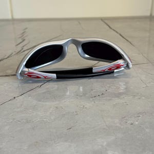 May include: Silver sunglasses with black lenses and red flame accents on the temples. The sunglasses have a sleek, modern design and are folded. The frame is silver and the lenses are dark.