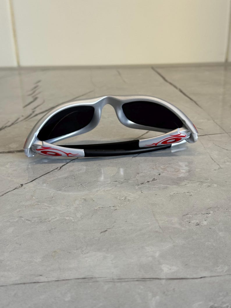 May include: Silver sunglasses with black lenses and red flame accents on the temples. The sunglasses have a sleek, modern design and are folded. The frame is silver and the lenses are dark.