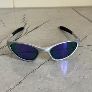 May include: Silver sunglasses with blue and green lenses. The sunglasses feature a curved design with black temple tips. The lenses reflect blue and green, offering a modern look. The frame is made of a light-colored metal.