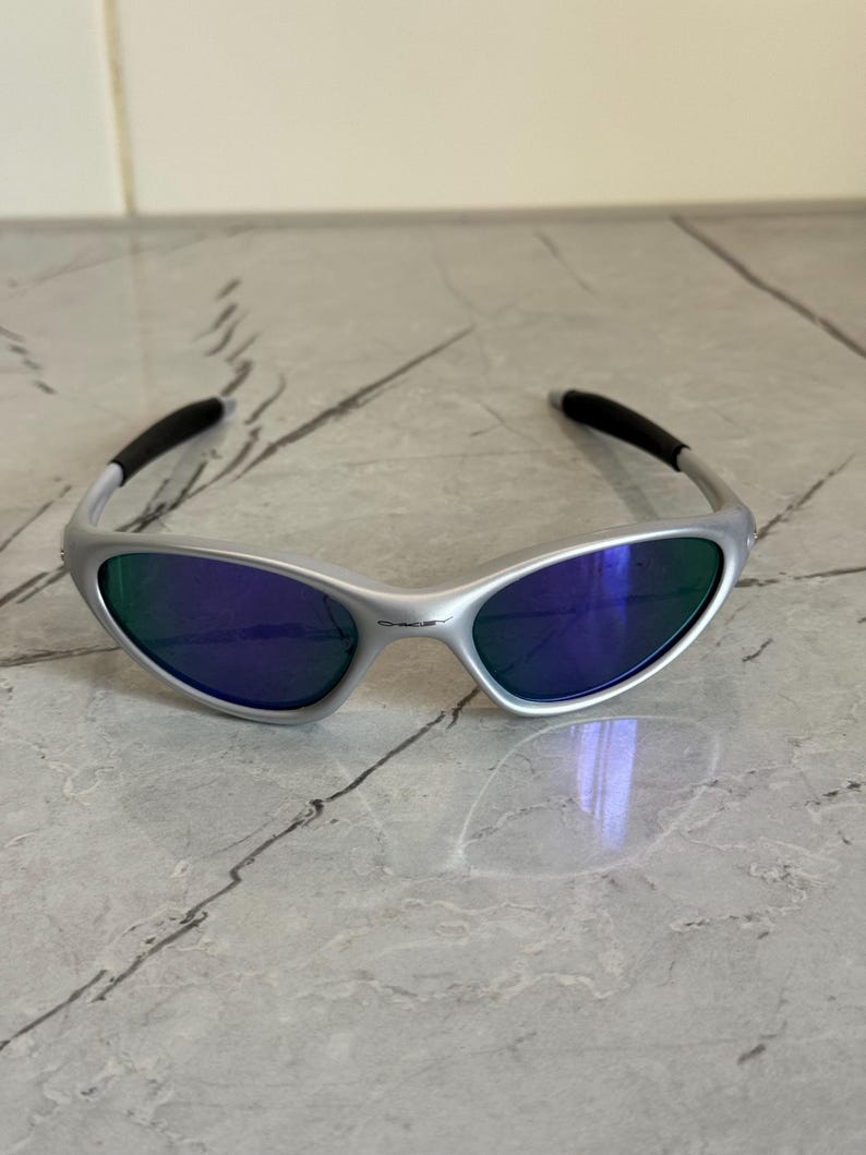 May include: Silver sunglasses with blue and green lenses. The sunglasses feature a curved design with black temple tips. The lenses reflect blue and green, offering a modern look. The frame is made of a light-colored metal.